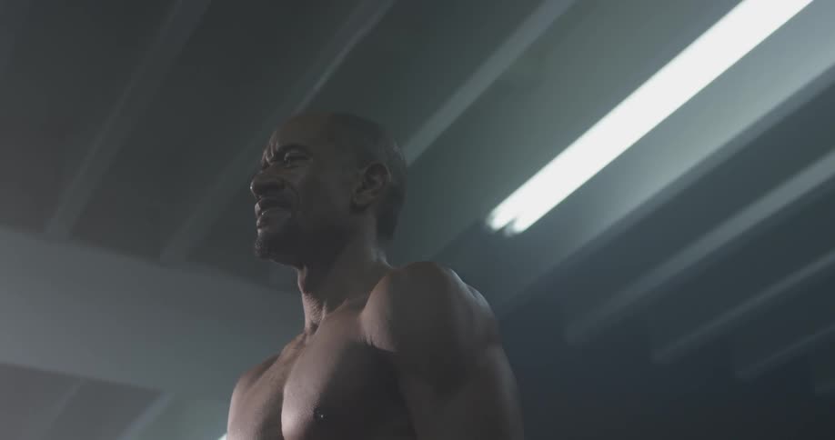 A muscular man grimaces as he stands under a fluorescent light