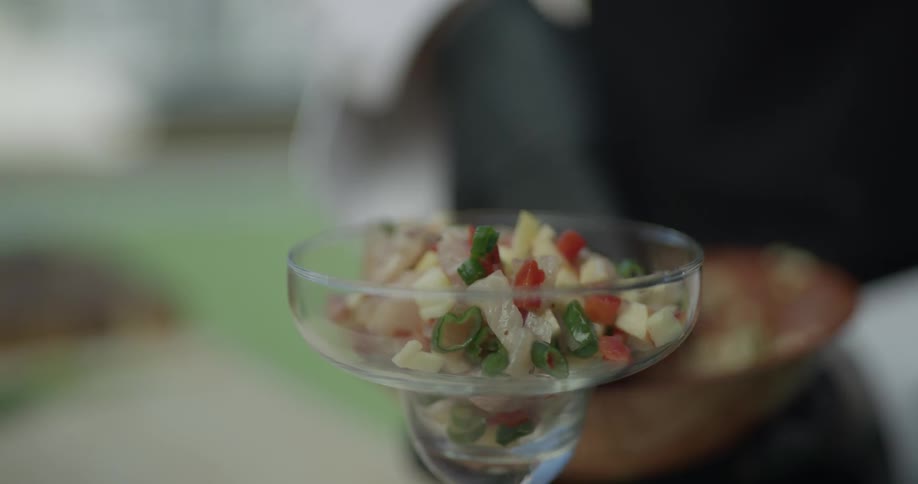 A chef places some vegetables inside a drinking glass fish too perhaps and wearing black gloves and black apron