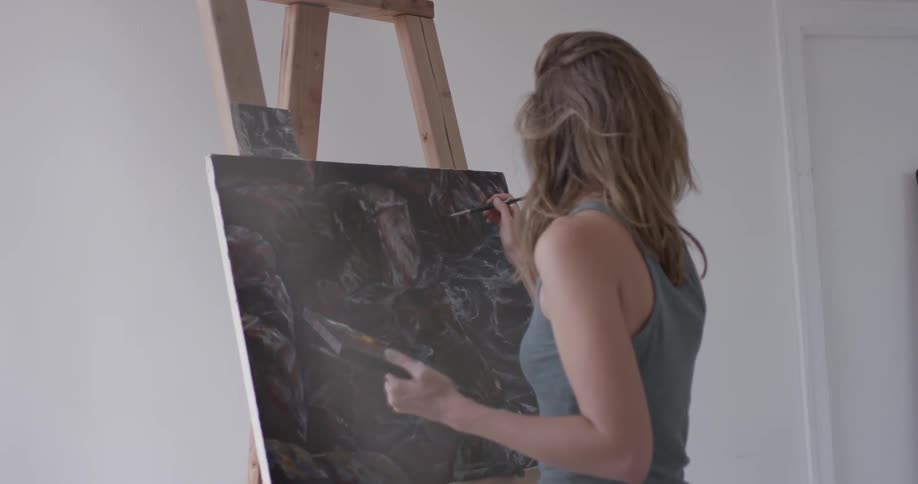 The top half of a woman in a gray tank top painting at an easel