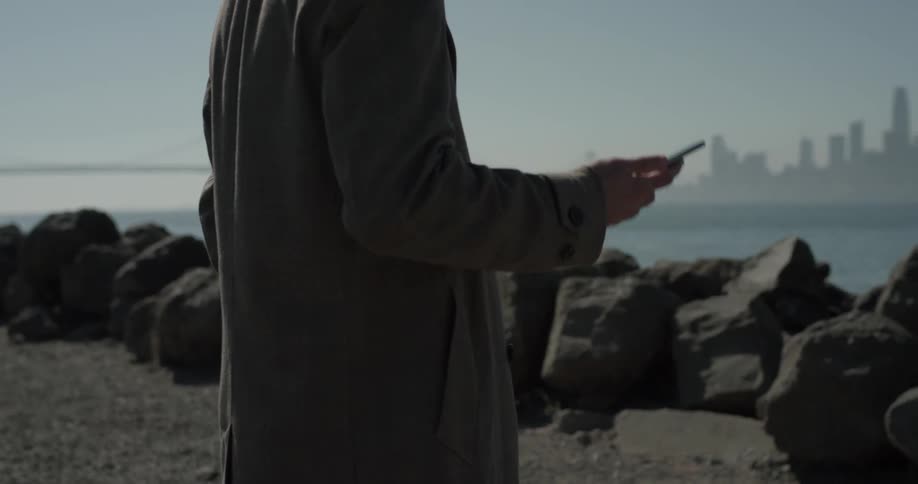 Man standing behind rocks near ocean taking out a cell phone