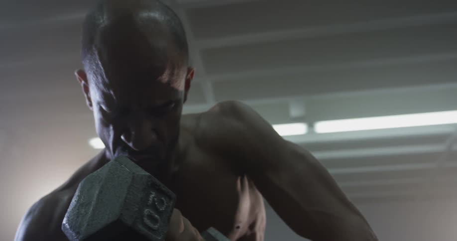 A male athlete is sitting in a gym in daylight doing curls with a weight
