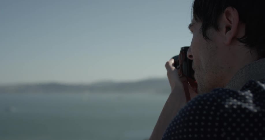 A man looks out over a large body of water probably a lake aiming his camera at the city on the horizon on a bright blue sunny day