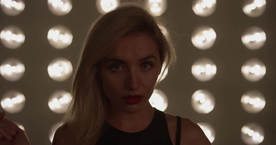 A woman touches her blond dyed hair wearing minimal clothing in front of a bunch of lights with red lipstick on in a dim lit room
