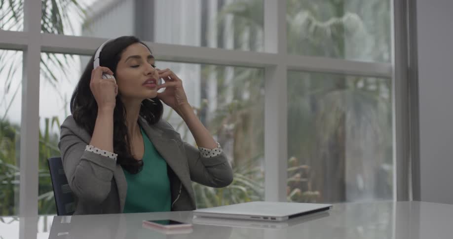 A woman sitting at a desk in daylight is closing her laptop and putting on earphones