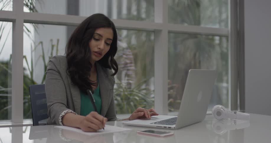 A woman wearing business attire sits at a desk working on a laptop and writing on a piece of paper