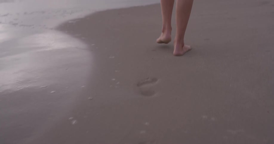 A caucAsian person walks through the beachfront as waves come in during the day time and footprints can be seen in the moist sand