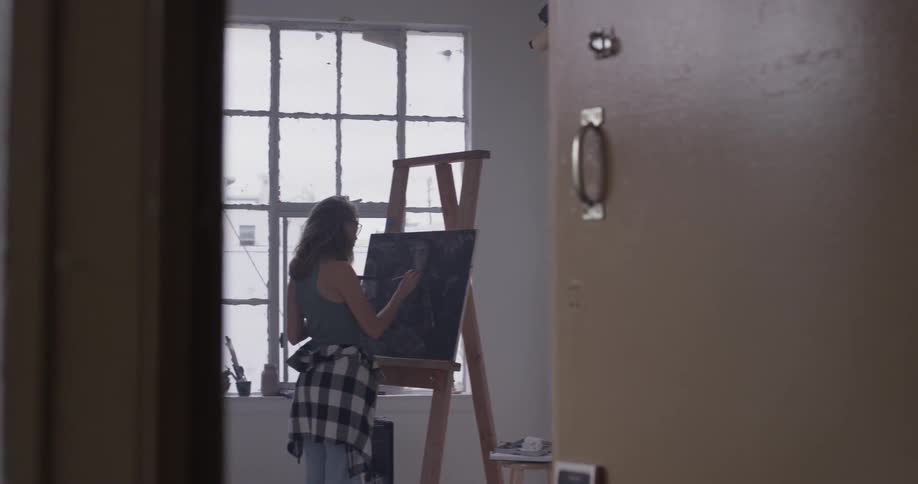A woman with a plaid shirt tied around her waist stands at an easel and paints