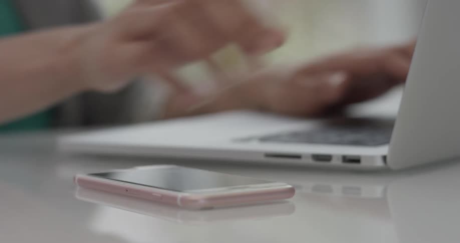 A person typing on a laptop picks up their cell phone