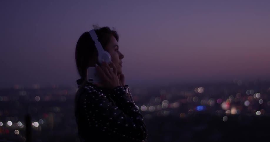 A woman stands overlooking the city after sunset wearing headphones and bobbing her head to music wearing a black leather jacket with metal studs