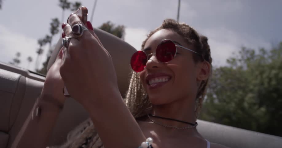 A woman wearing sunglasses yells and takes pictures with her phone while riding in the back of a convertible