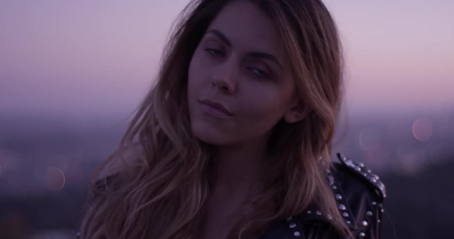 Sexy beautiful long hair gorgeous woman with leather metal studded black jacket stands and crosses her arms with the sunset behind her
