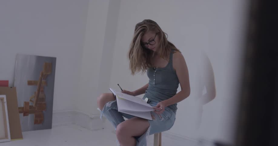 Woman sits on a stool in an empty room looking down at a notebook