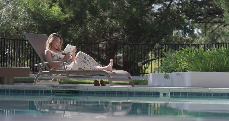 A woman lounges in a pool chair by the pool reading a book