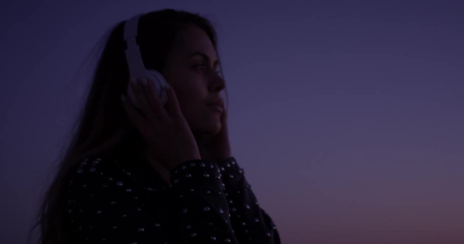 Woman stands outside at sunset listening to headphones