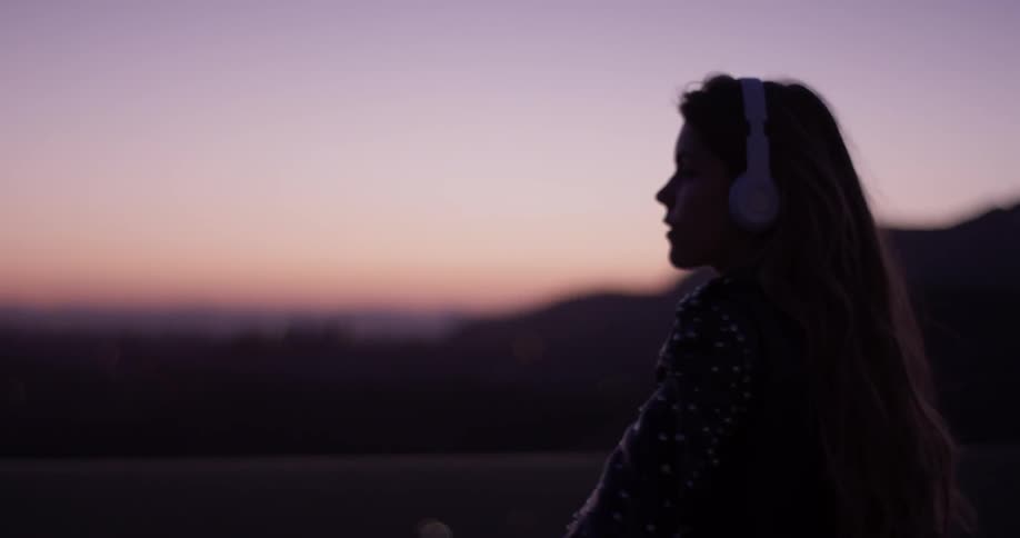 Woman wearing earphones stands outside at sunset