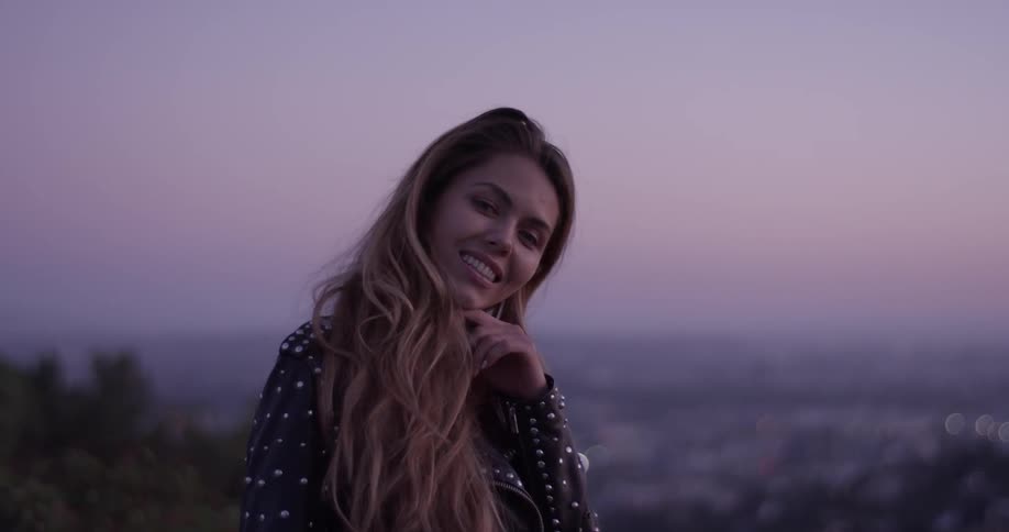 Women who is beautiful has long hair and a leather jacket with metal studs stands on the hilltop overlooking a city