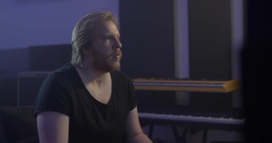 A man sitting in a music studio surrounded by keyboards is bobbing his head