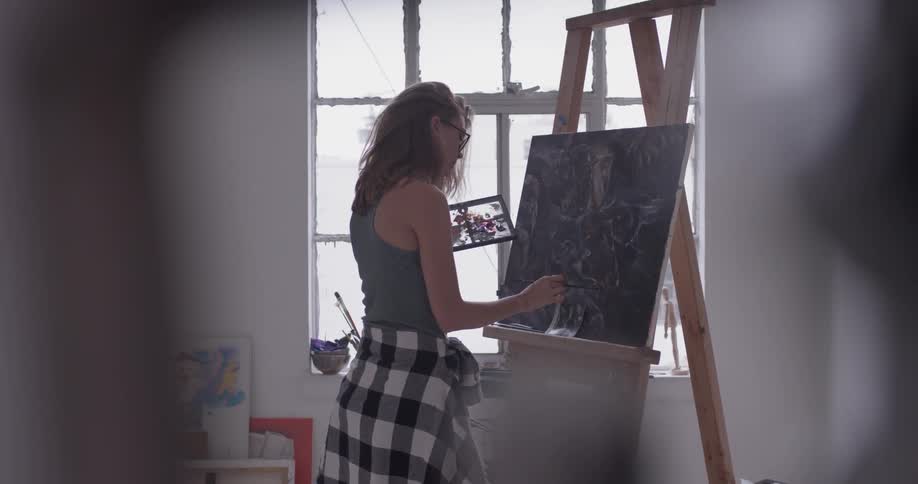Woman wearing a tank top stands in front of a window and paints at an easel