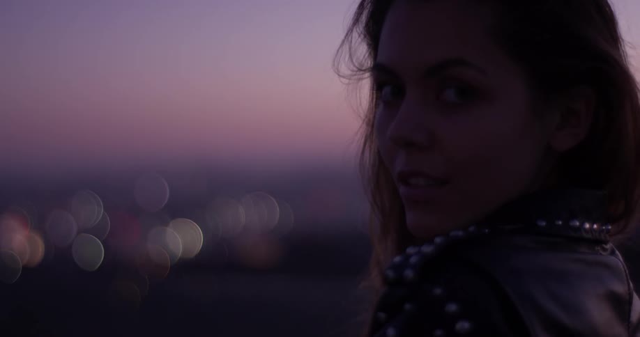 A closeup of a woman turning her head and looking sultry with a lit up city in the background