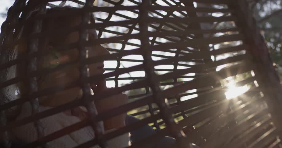 A woman sits in an egg shaped rattan chair outside as the sun goes down