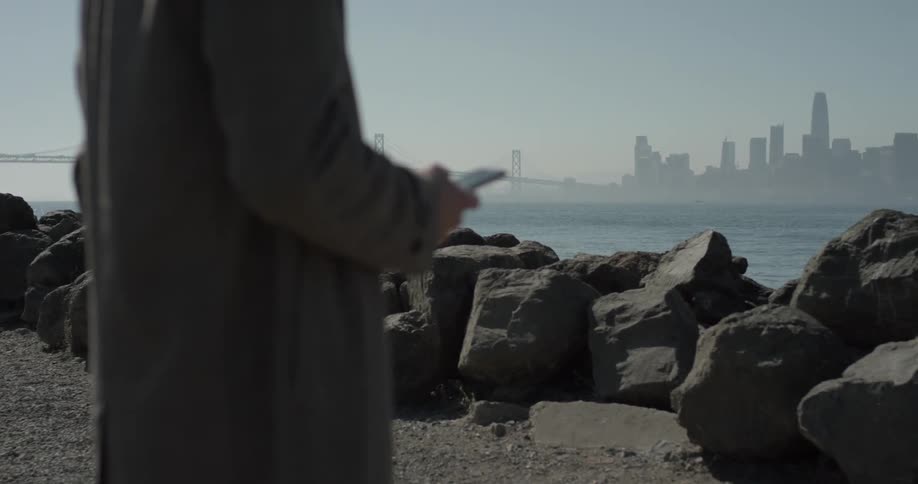 A man stands by some rocks at the shoreline of the great body of water with a large city on the horizon of a bright sunny day