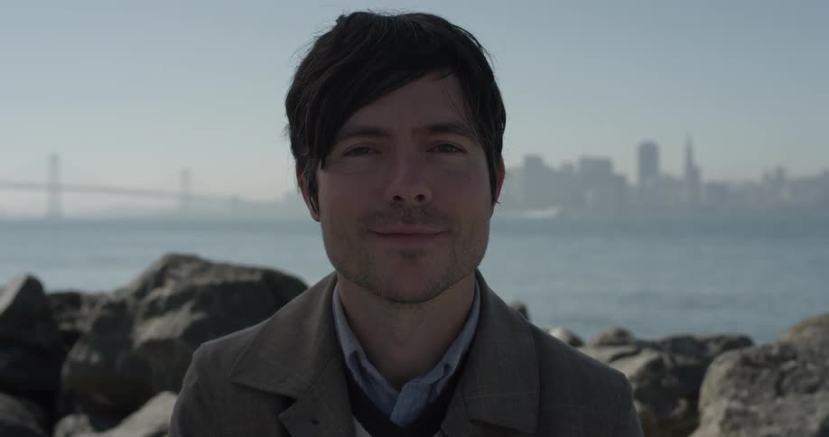 Man looks at camera with rocks, water, bridge, and city in background