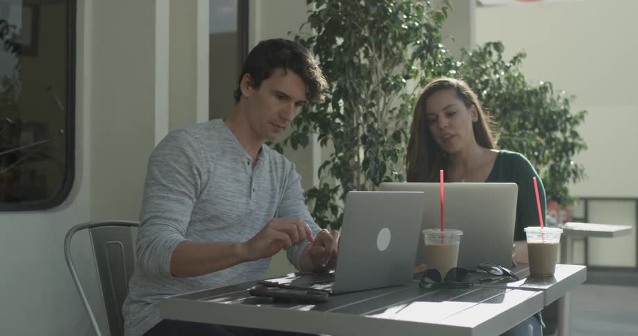 A man and women are at a coffee shop having coffee and chatting about things on their laptops on the table on a bright sunny day
