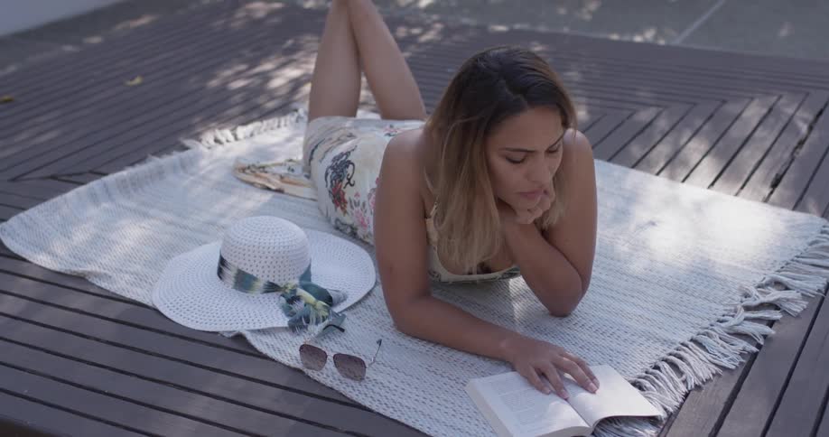 A woman lays on her stomach on a deck reading a book