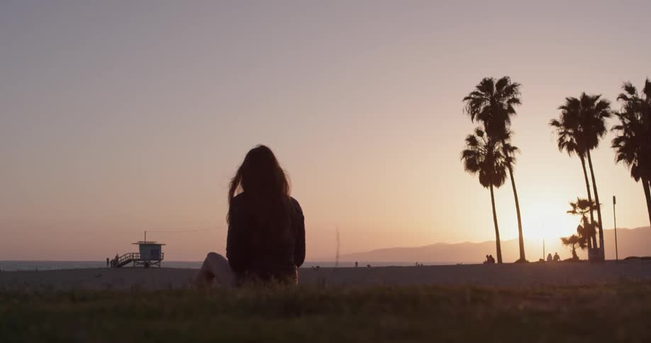 A woman with long hair sits on the grass looking at the beach at sunset