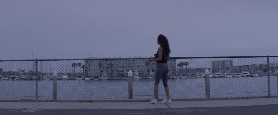 A woman jogs over to a fence by the water and stretches her legs