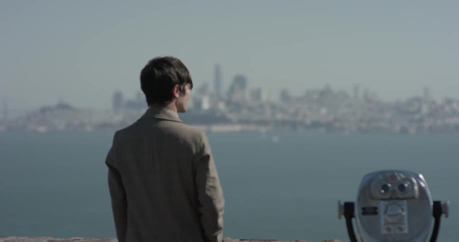 A man stands from an observation area standing and staring at a major city on the horizon over a body of water on a bright sunny day