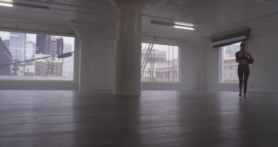 A woman with a dancers figure walks on a wooden floor in an empty room