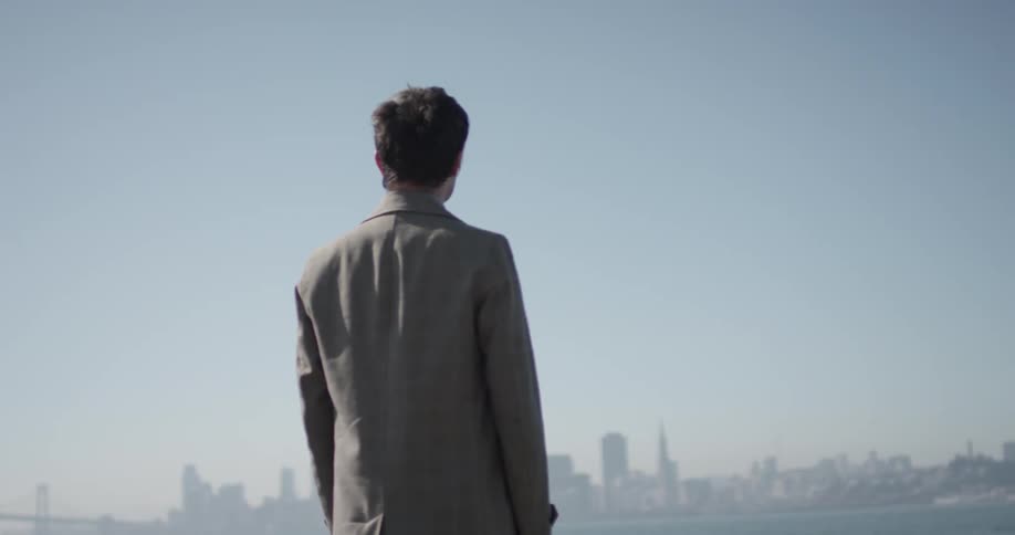 Man stands on top of a rock overlooking a great body of water with a large city off in the horizon of a bright sunny blue sky day