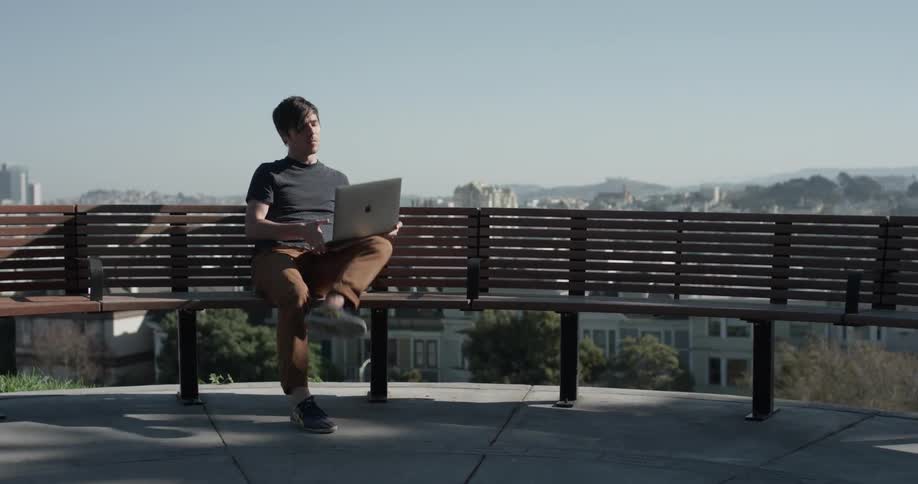 Man sits on a bench in the city looking at his camera and picks up a laptop