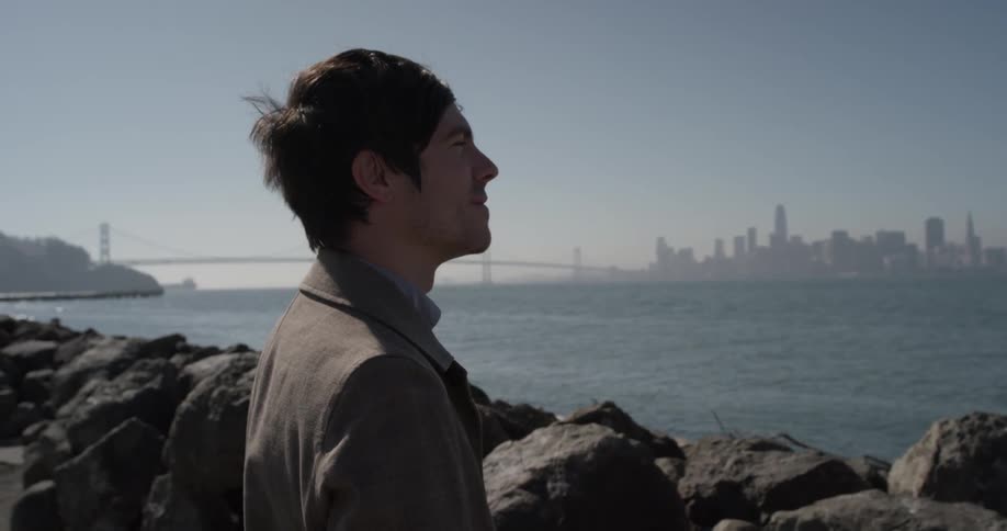 A caucasian man stands next to large boulders at the edge of the ocean with a city in the background
