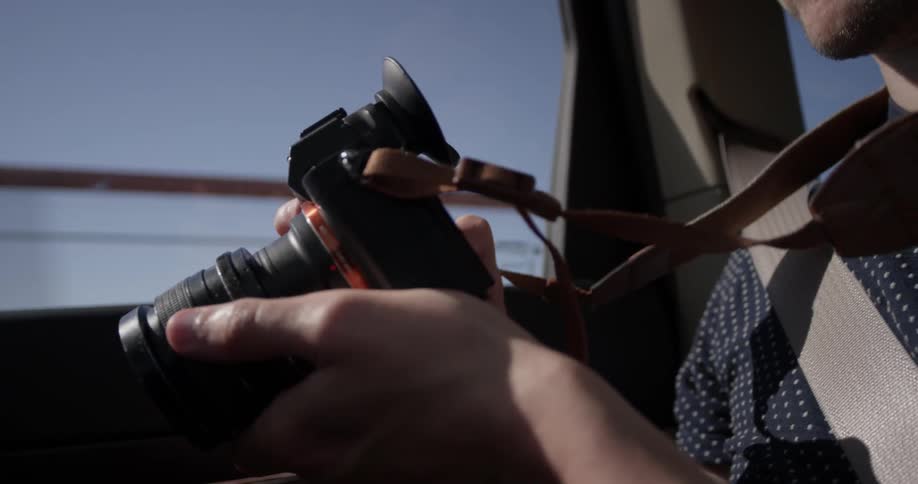 A man holding a camera walks drives over a bridge in the passenger seat holding his camera on a bright blue sunny day