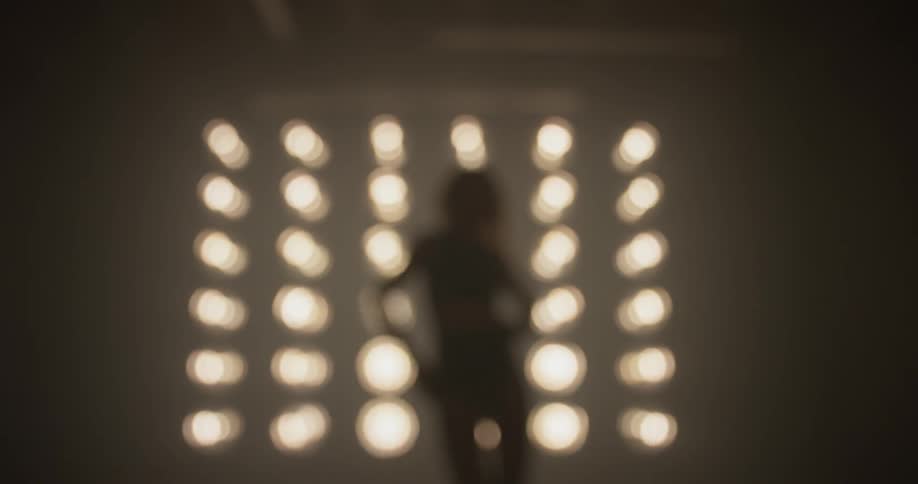 Women dances sexually in front of a grid of bright lights with black underwear on in a dim lit room