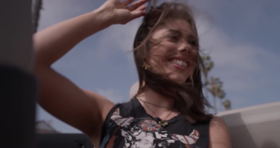 Woman dances to music with the top off of the car and sunglasses on head with wind in her hair on a bright sunny day