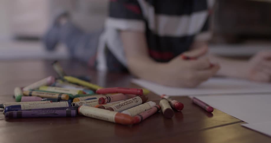 Many crayons are on the floor in front of a child who is drawing and he picks a new one to continue sketching his sheet of paper