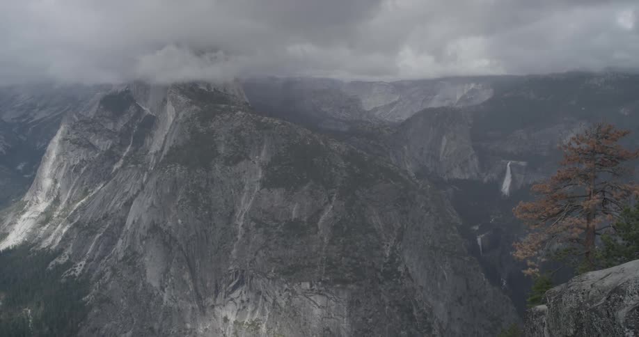 An aerial view of a mountain range with fog and clouds rolling over while waterfalls pour off the cliffs and trees adorn