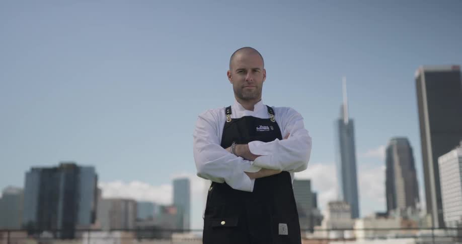 A white man with extremely short hair stands with a black apron up top of a building with the city skyline behind him