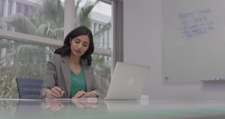 Woman looks at whiteboard, types on laptop, and writes in notebook