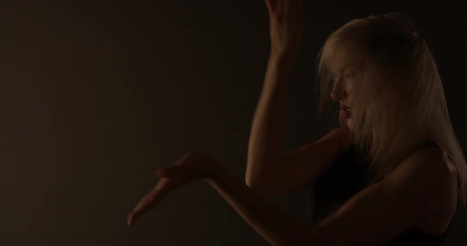 A fake blond women with makeup and minimal black clothing dances enticingly and passionately inside a dimly lit room with lots of aesthetically pleasing lights