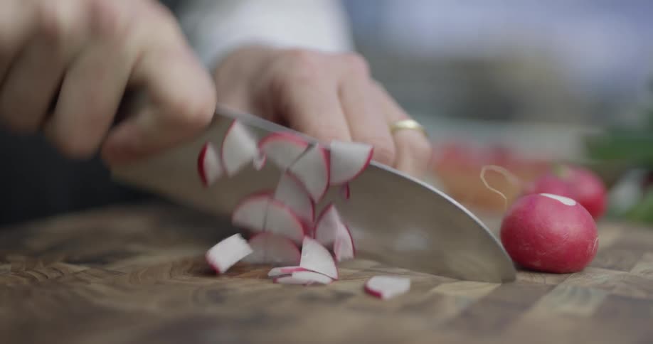 Radishes are cut on a cutting board and scooped up with a knife by the chef wearing a black apron