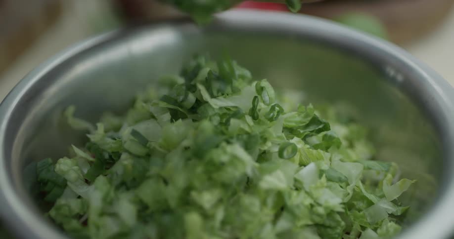 Lettuce and tomatoes inside a metallic bowl have fresh chives placed over the top by a chef with a sharp knife