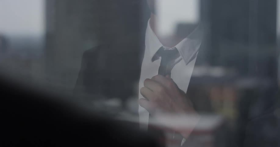 A man adjusts his tie for a suit he is wearing in the reflection of a tall window which overlooks the city ahead