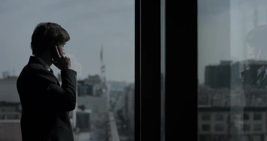 A man talks on the phone as he stands in front of a window overlooking a city