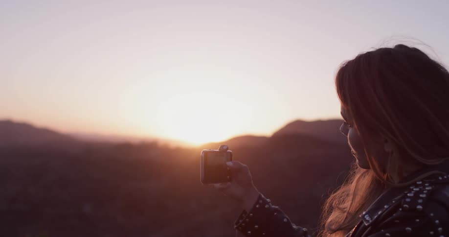 A woman is taking a selfie on top of a hill with her camera phone as the sun sets in front of her