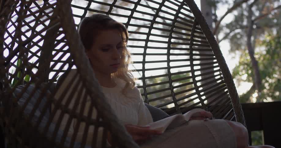 A woman sits in a rattan swing reading a book