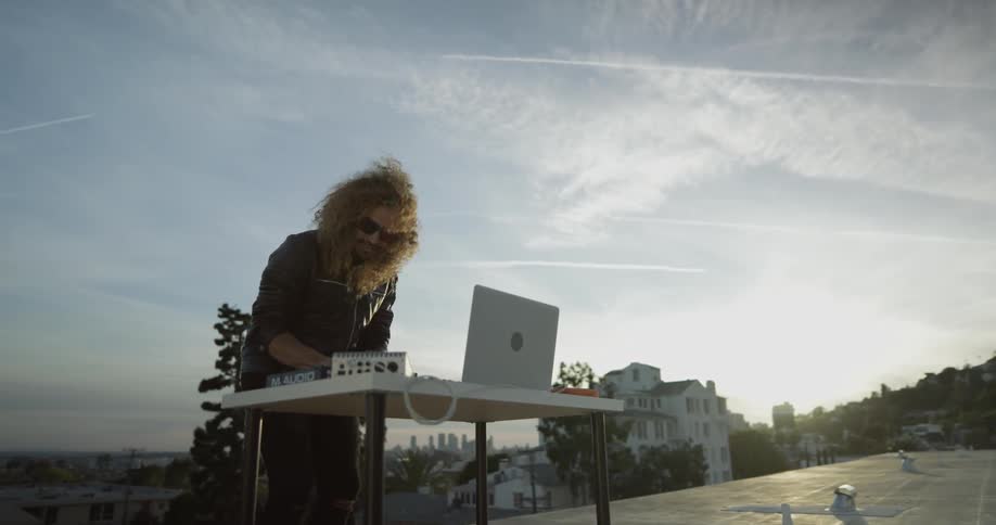 A man stands behind a small white table with a laptop on it and other dj equipment banging his head playing some music
